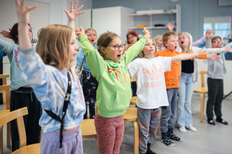 Lapset venyttelevät käsiä seisoen ja laulavat samaan aikaan. Kuoron äänenavaushetki.